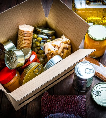 High angle view of a cardboard box filled with multicolored non-perishable canned goods, conserves, sauces and oils shot on wooden table. The composition includes cooking oil bottle, pasta, crackers, preserves and tins. High resolution 42Mp studio digital capture taken with SONY A7rII and Zeiss Batis 40mm F2.0 CF lens